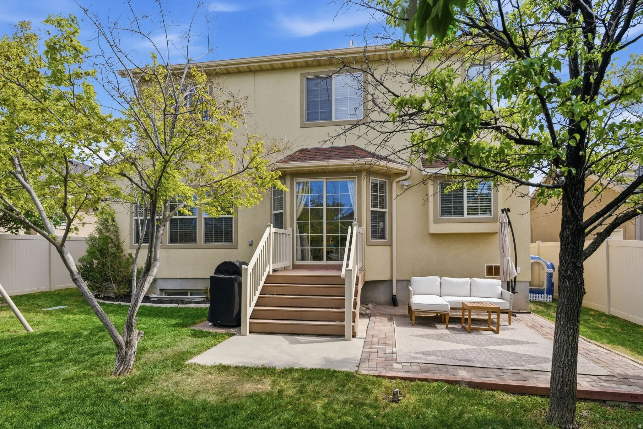 Rear exterior featuring a spacious green lawn, a multi-level deck with stairs, a brick paver patio, a bay window, and mature trees providing natural shade
