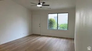 Spacious room with wood-finish flooring, a contemporary ceiling fan with integrated lighting, a large window, white walls, and a white interior door