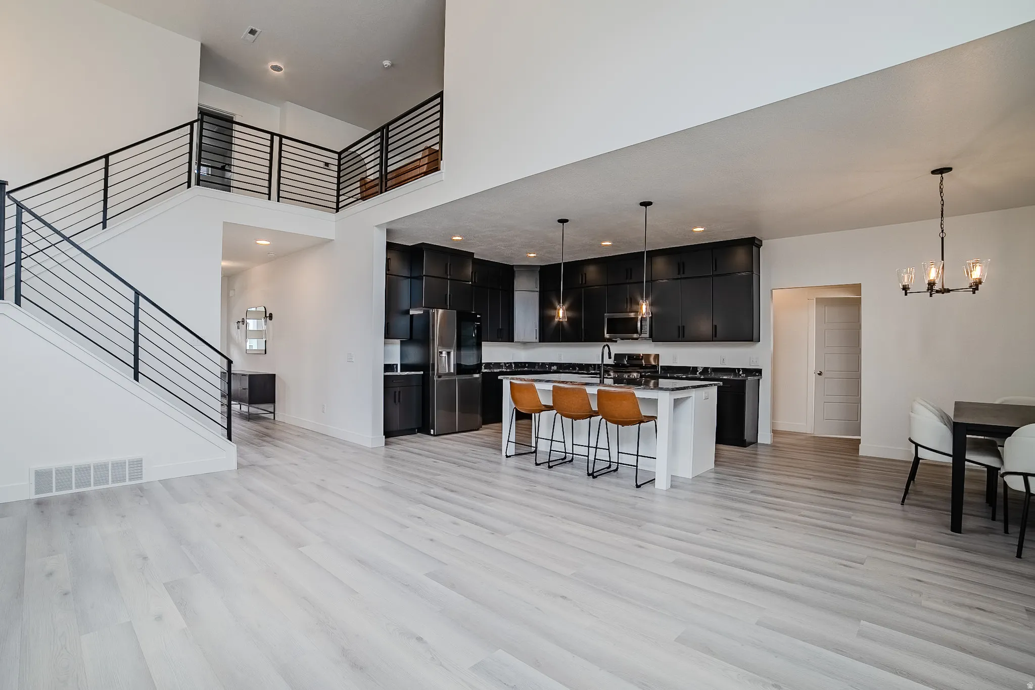 Open-concept living area featuring light wood-finish flooring, a contemporary kitchen with dark cabinetry and stainless steel appliances, a spacious central island, a dining area with modern chandelier, and a two-story ceiling