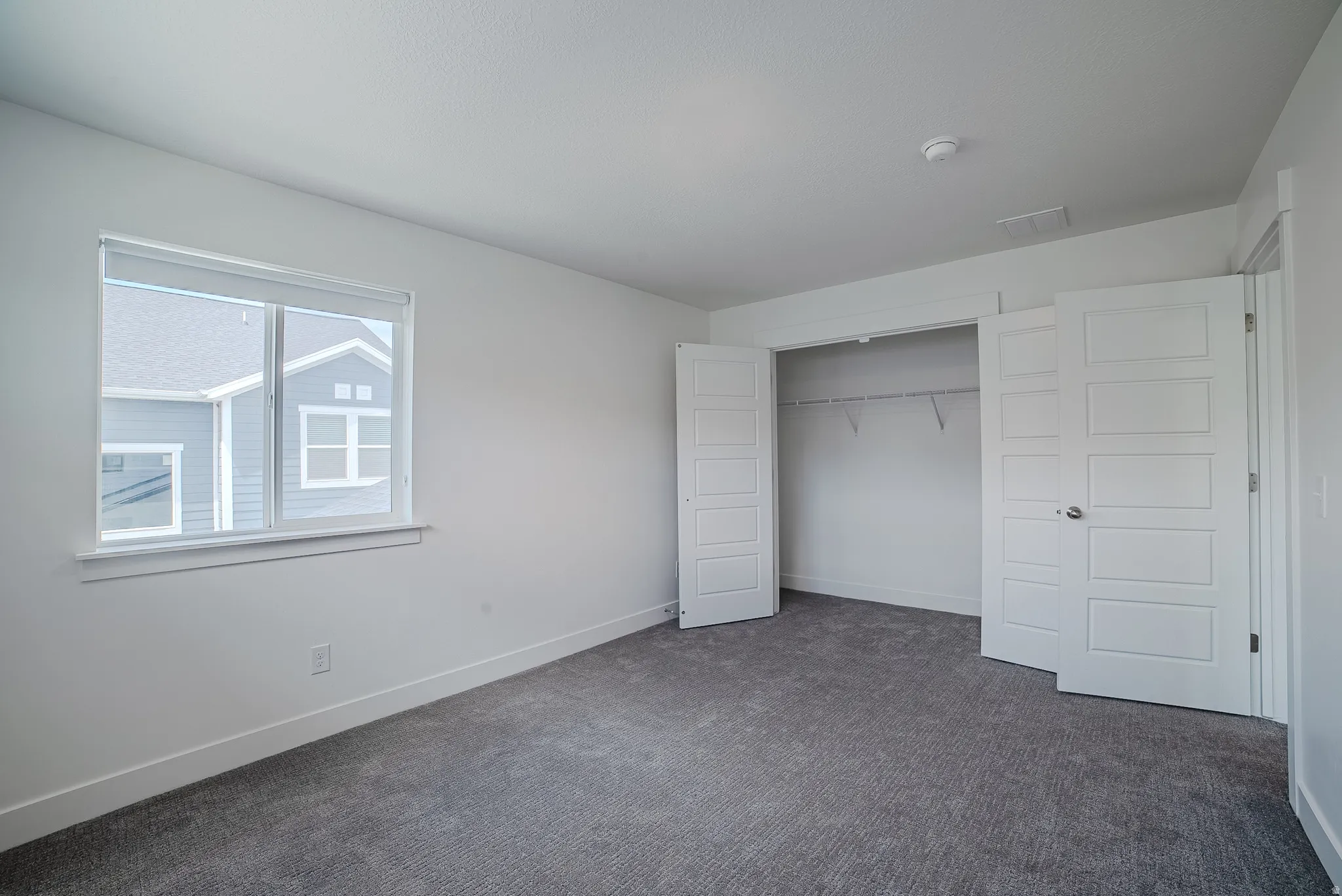 Spacious room featuring neutral carpeting, a large window with roller shades, and a built-in closet with bifold doors