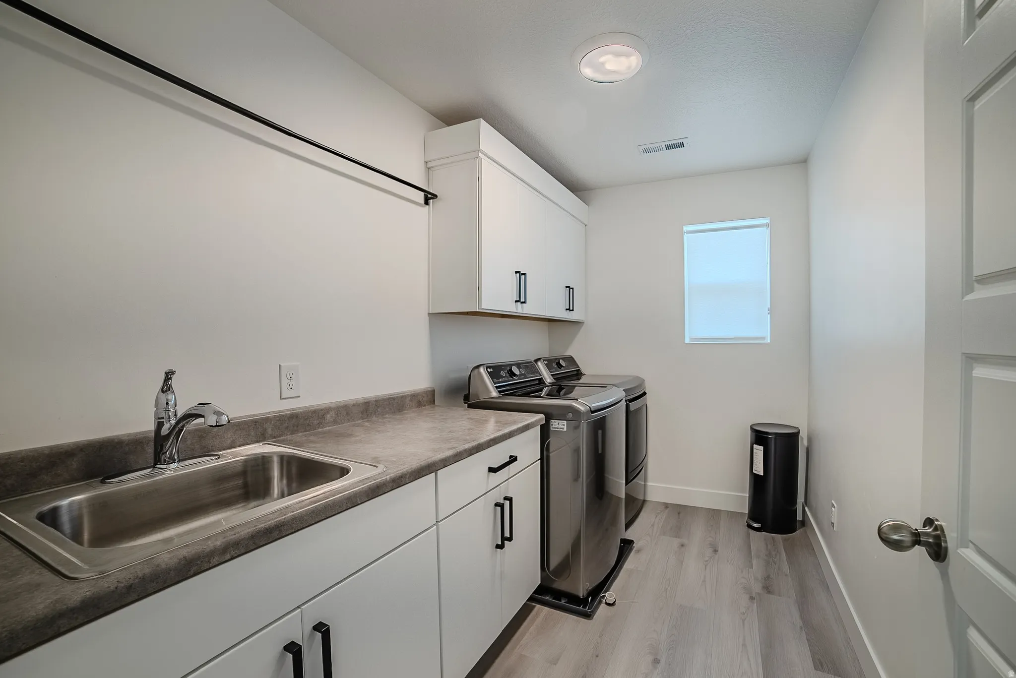 Dedicated laundry room featuring light wood-finish flooring, white cabinetry with black hardware, a stainless steel sink, a gray countertop, and a window for natural light