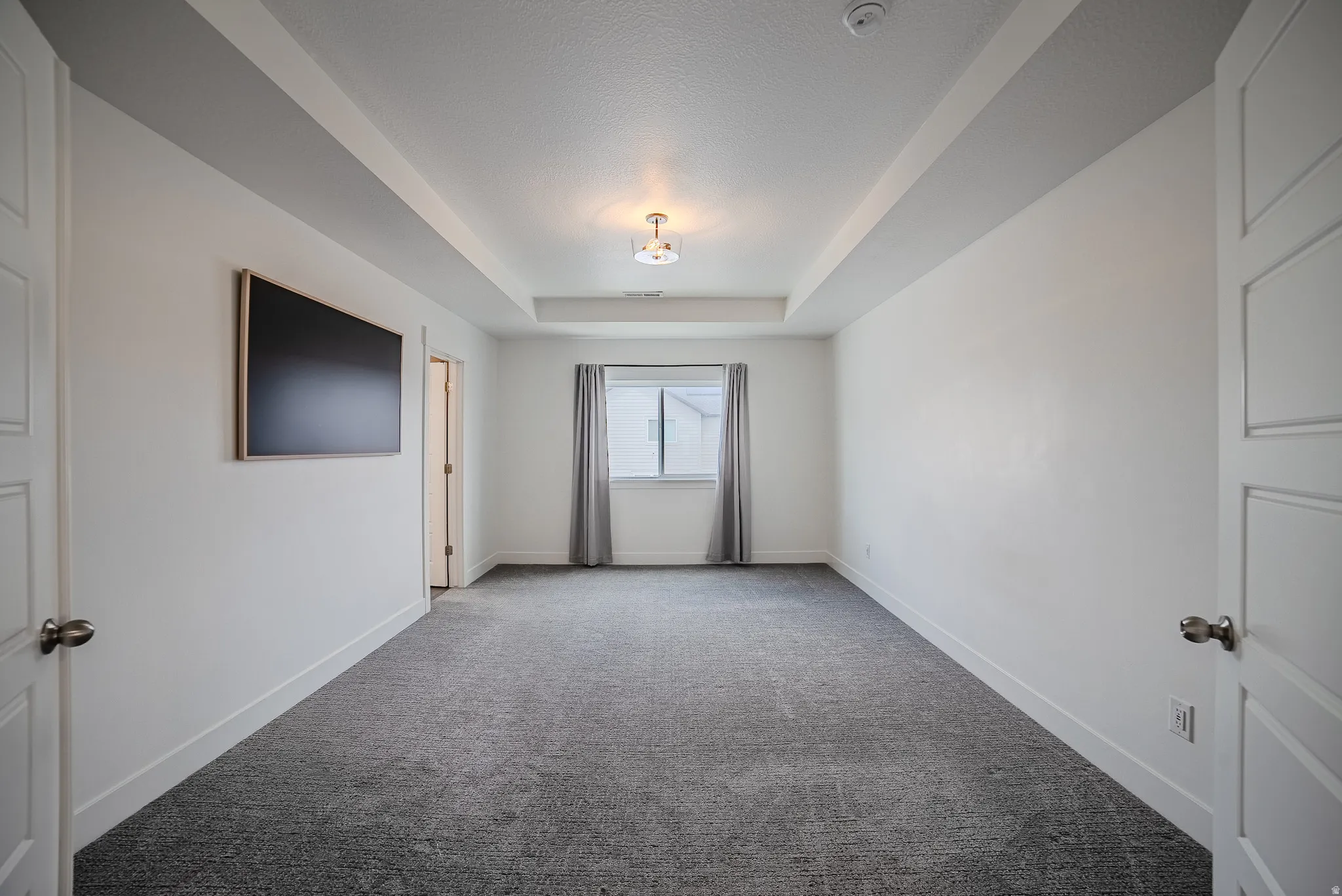 Spacious room featuring a tray ceiling with recessed lighting, plush gray carpet, and a large window with gray drapes