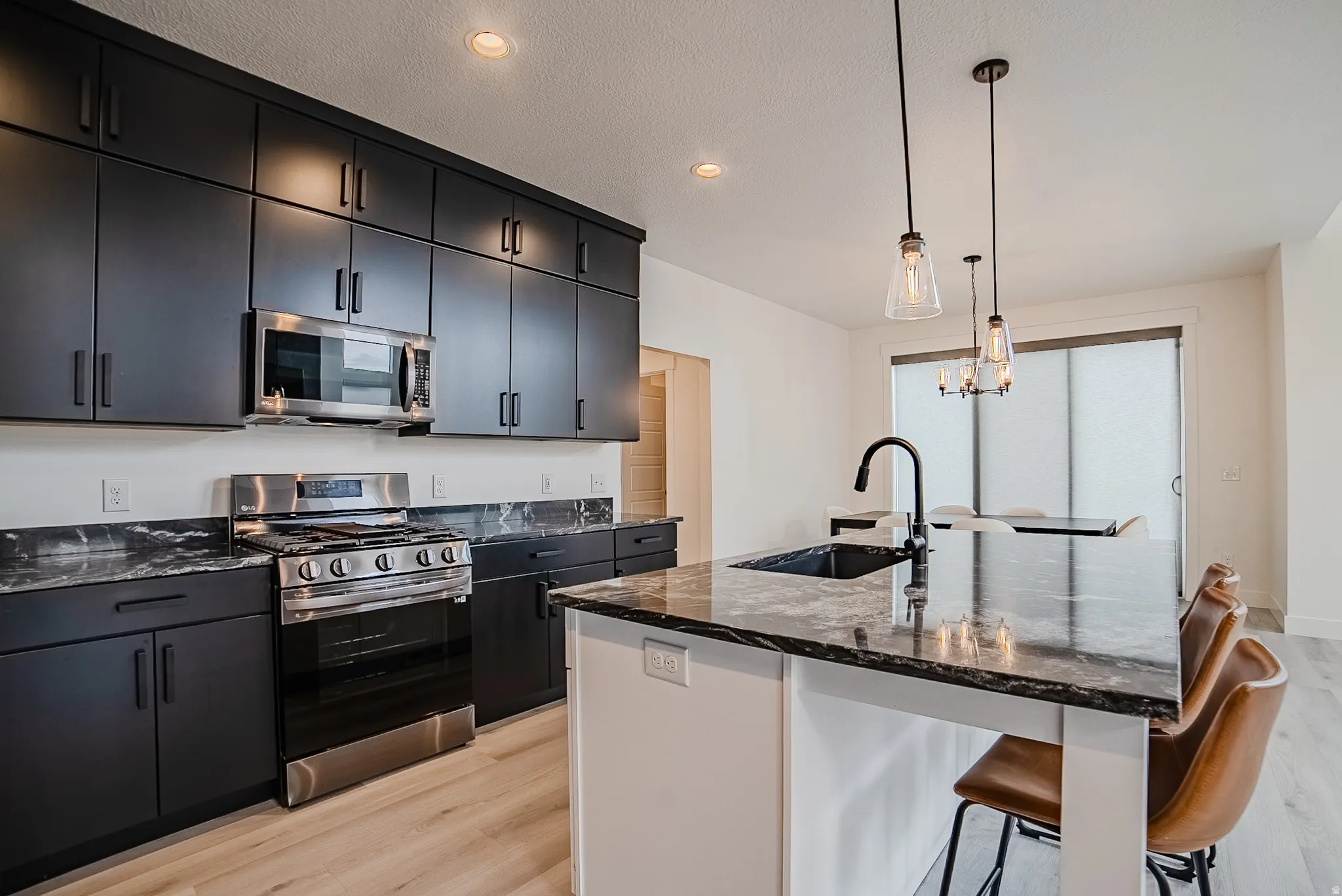Modern kitchen featuring dark cabinetry, stainless steel appliances, dark stone countertops, a large island with an integrated sink, and pendant lighting