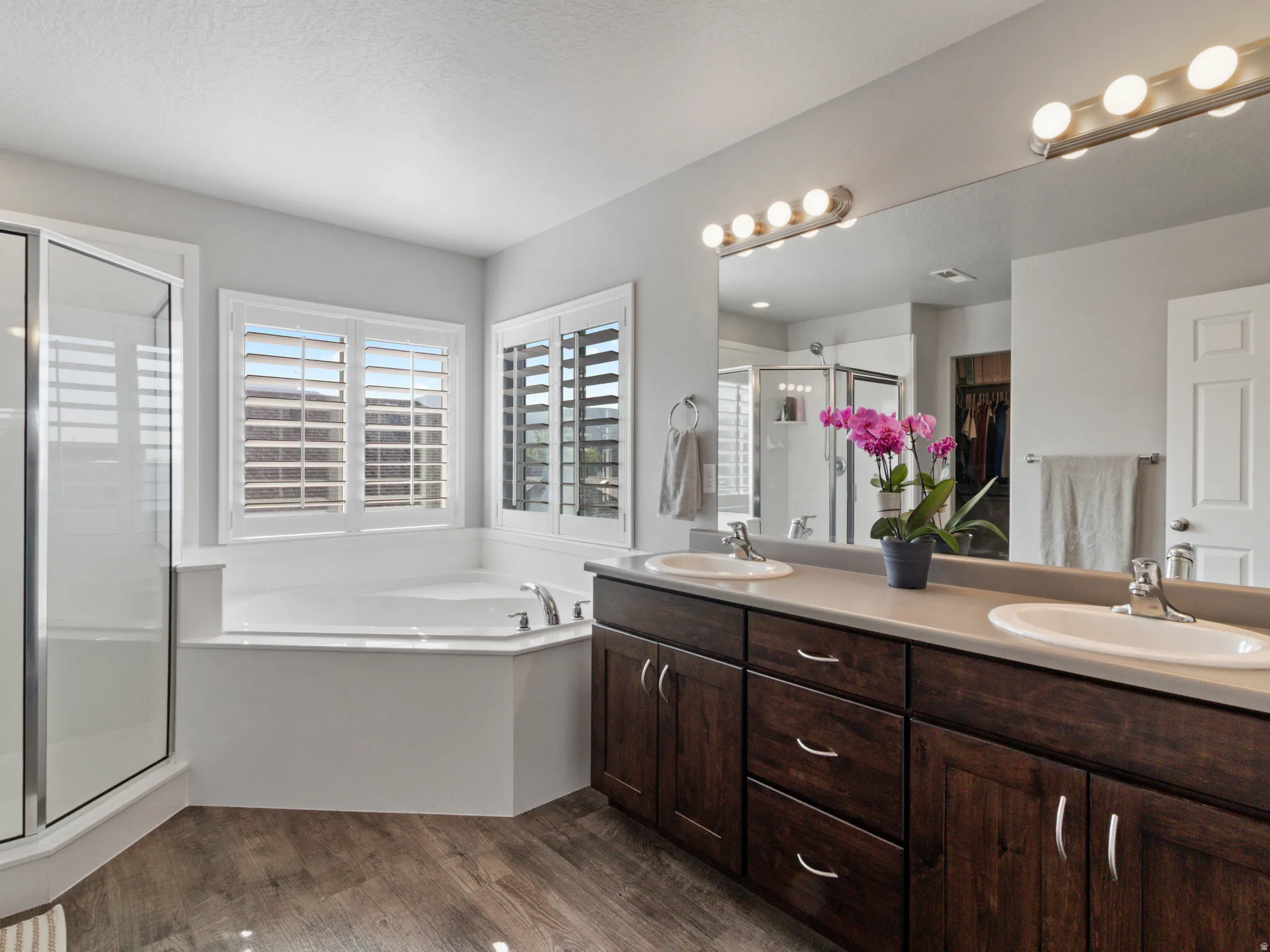 Luxurious bathroom featuring a corner soaking tub, separate glass-enclosed shower, dual vanities with wood-finish cabinetry, and wood-finish flooring