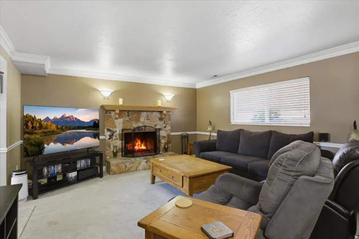 Stone surround fireplace with hearth, wall sconce lighting, window with blinds, crown molding, and carpeted flooring