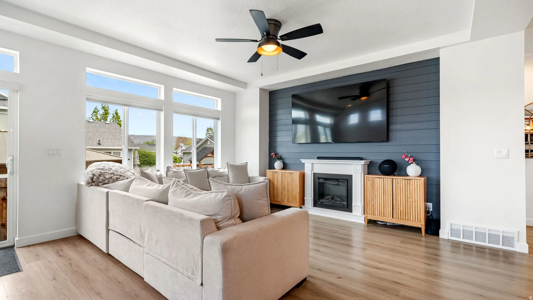 Spacious living area featuring large windows, wood-finish flooring, a ceiling fan, and a fireplace with a white mantel and a shiplap accent wall