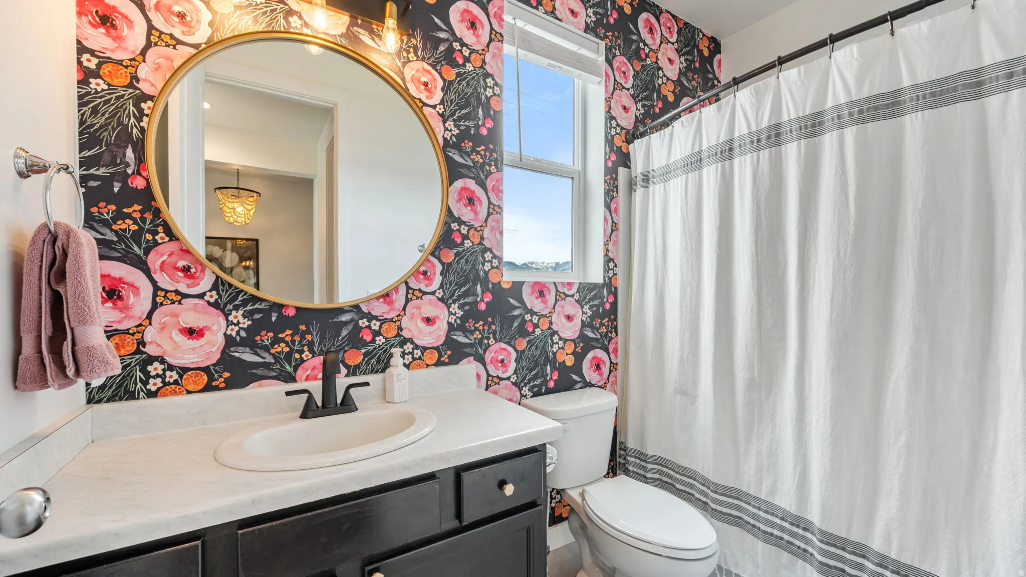 Bathroom featuring floral wallpaper, a dark wood vanity with a light countertop, a round gold-framed mirror, and a window providing natural illumination