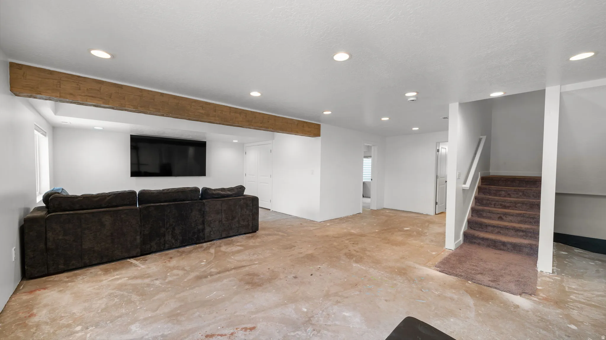 Spacious lower-level area featuring a rustic wood beam, recessed lighting, and a white painted staircase with a carpeted runner
