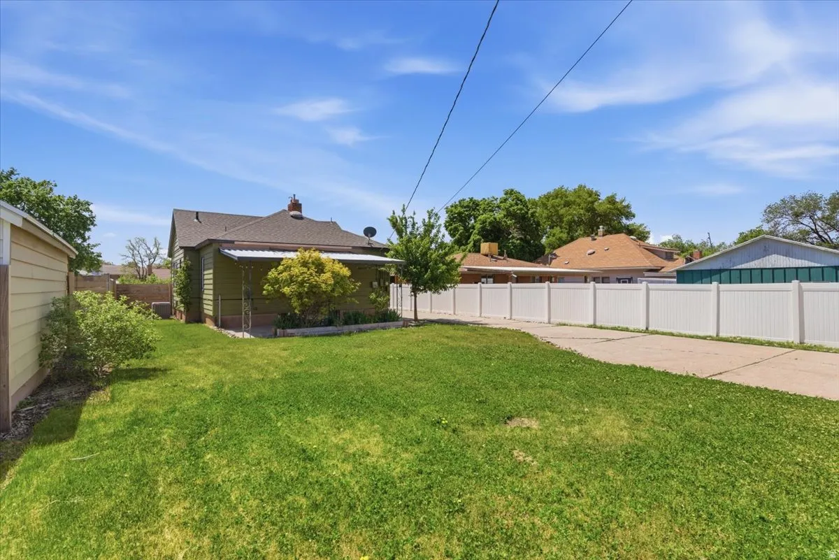 Expansive backyard featuring a green lawn, a concrete driveway, a white privacy fence, a detached shed with horizontal siding, and mature trees