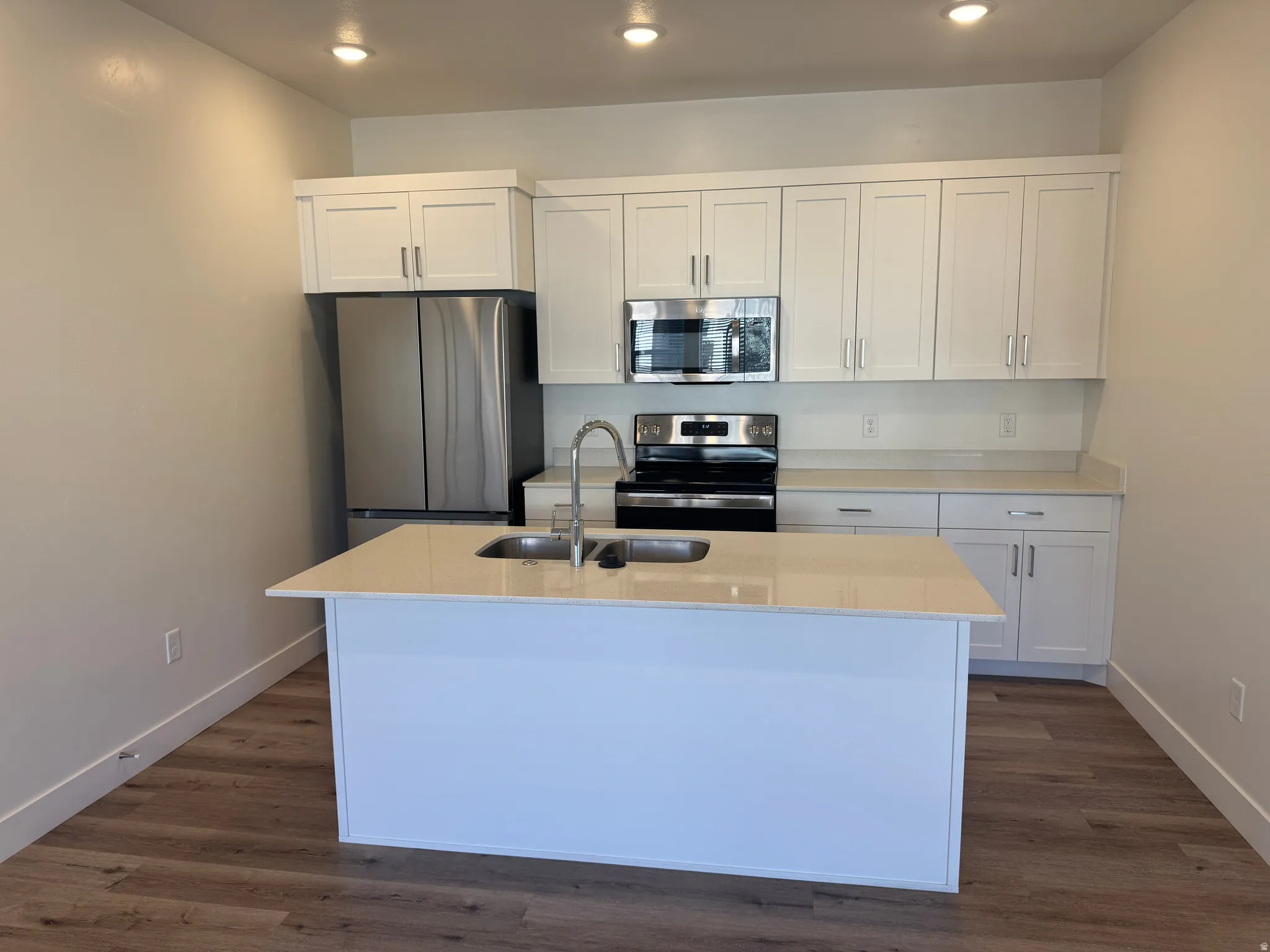 Modern kitchen featuring a central island with an undermount double sink and chrome faucet, white cabinetry, light-toned countertops, stainless steel appliances, and wood-finish flooring
