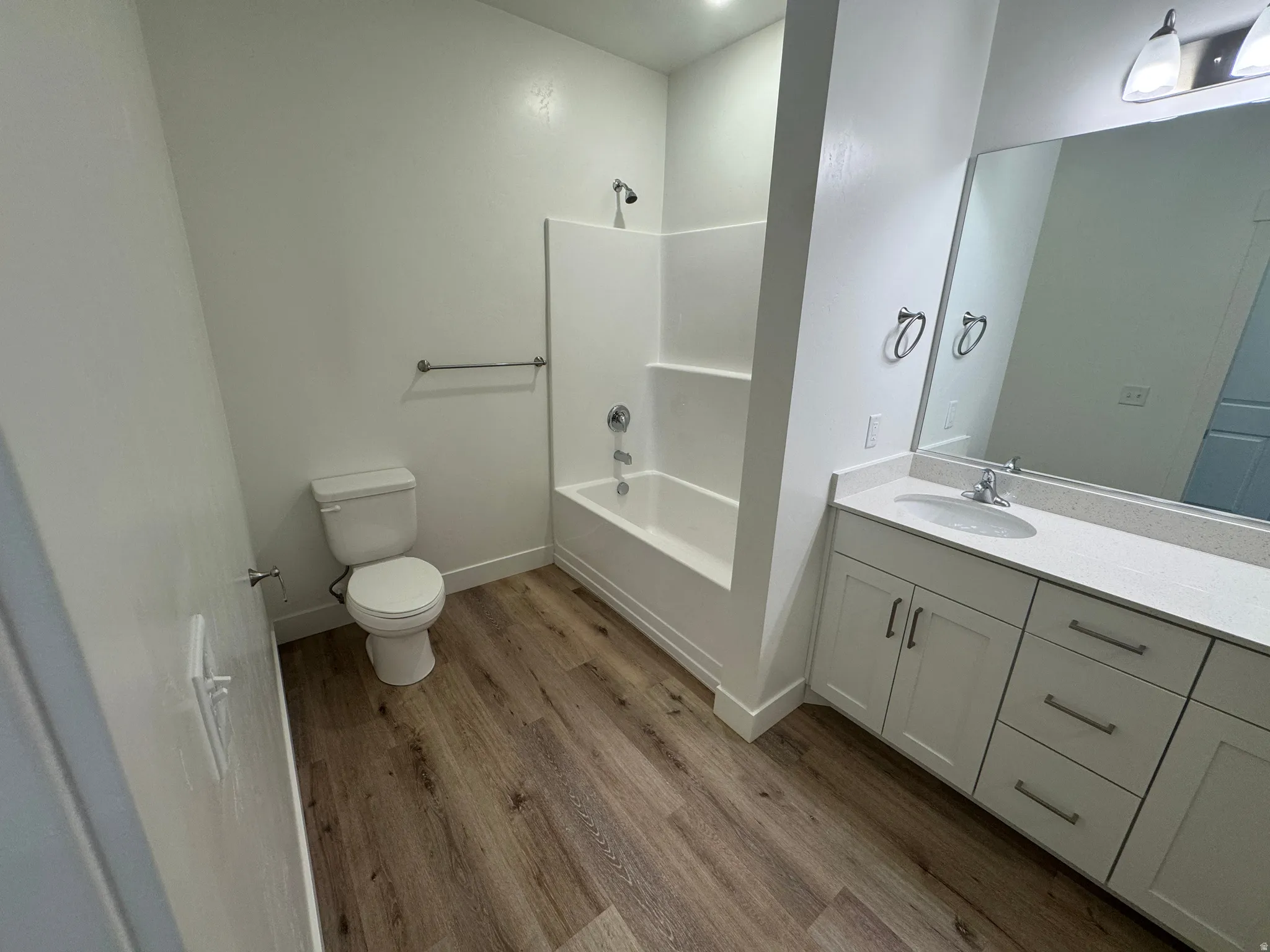 Bathroom featuring wood-finish flooring, a white vanity with shaker-style cabinetry, a built-in tub and shower combination, a white toilet, and a large wall-mounted mirror