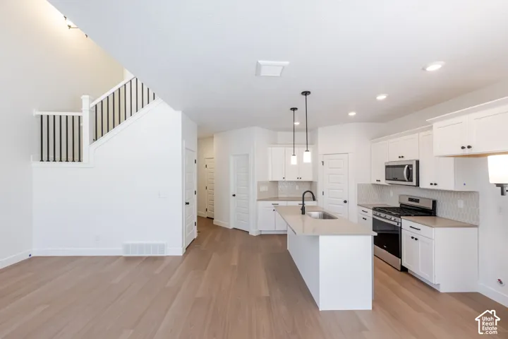 Open-concept design featuring wood-finish flooring, a contemporary kitchen with white cabinetry, a central island with a sink, stainless steel appliances, and a staircase with black metal balusters