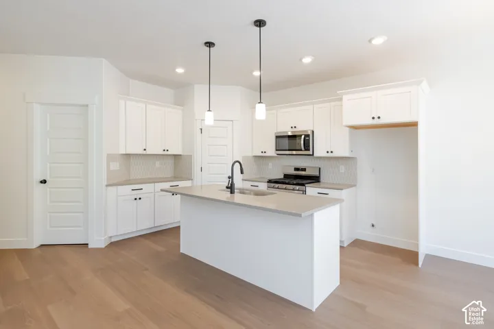Kitchen featuring white cabinetry, light-toned countertops, a central island with sink, stainless steel appliances, and wood-finish flooring