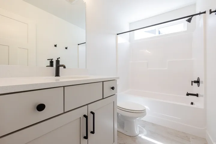 White bathroom featuring a single vanity with stone-finish countertop, matte black fixtures, a built-in tub with a shower, and a high-set horizontal window