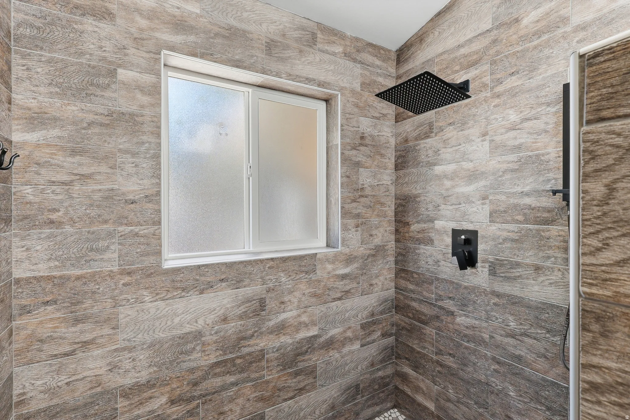 Shower enclosure featuring wood-grain tile walls, a frosted window, and matte black fixtures including a rain shower head and integrated controls