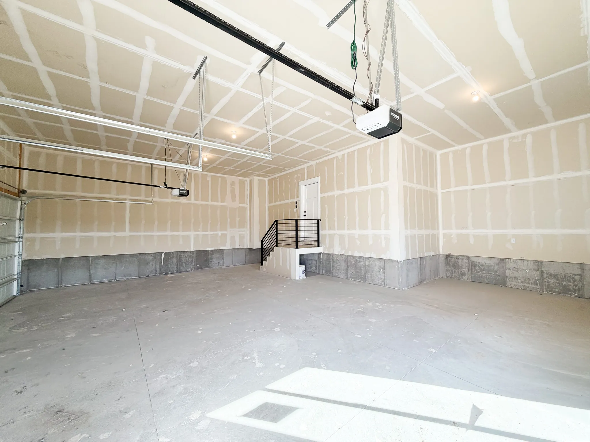 Spacious garage featuring concrete flooring, unpainted drywall, exposed ceiling joists, and a built-in staircase with a black metal handrail leading to a white interior door