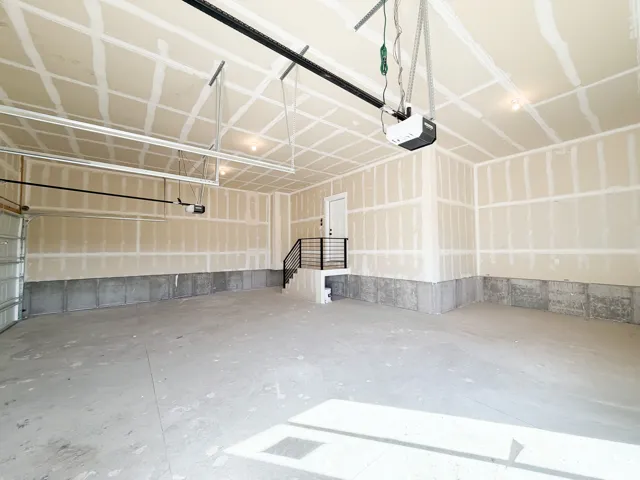 Spacious garage featuring concrete flooring, unpainted drywall, exposed ceiling joists, and a built-in staircase with a black metal handrail leading to a white interior door