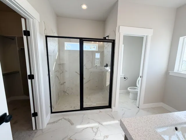 Spacious bathroom featuring a frameless glass shower enclosure with matte black accents, marble-look tile flooring, and a stone-finish countertop with an integrated sink