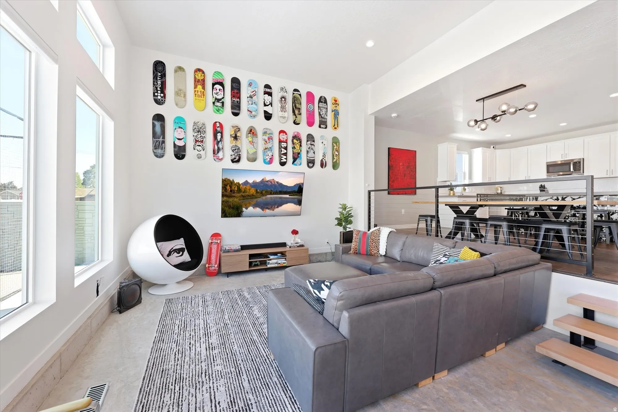 Open-concept living area featuring multiple large windows, a raised kitchen with pendant lighting, a black metal railing, and exposed wood stair treads