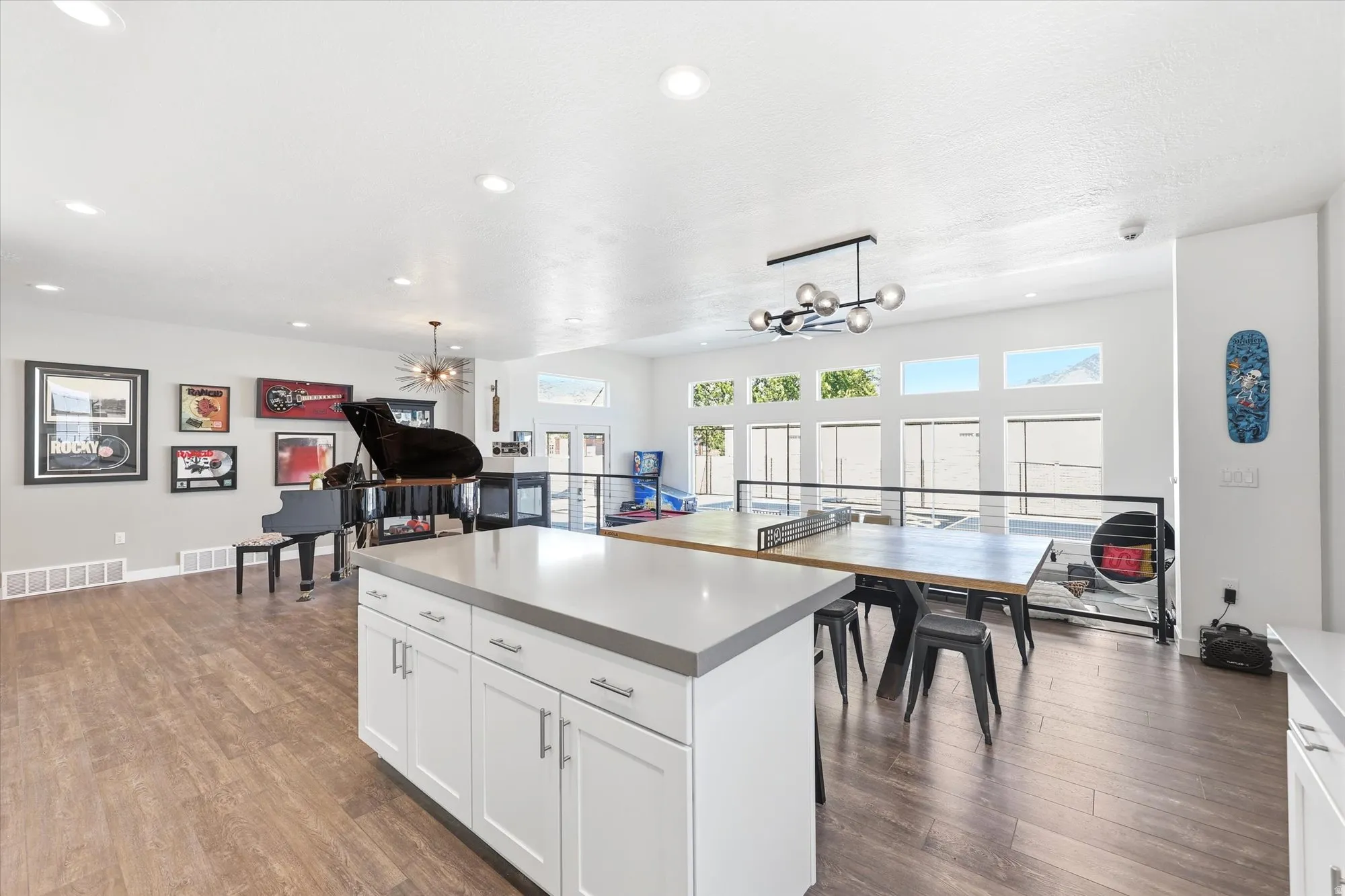 Open-concept area featuring wood-finish flooring, a white kitchen island with a light-toned countertop, recessed lighting, and large windows with transom details