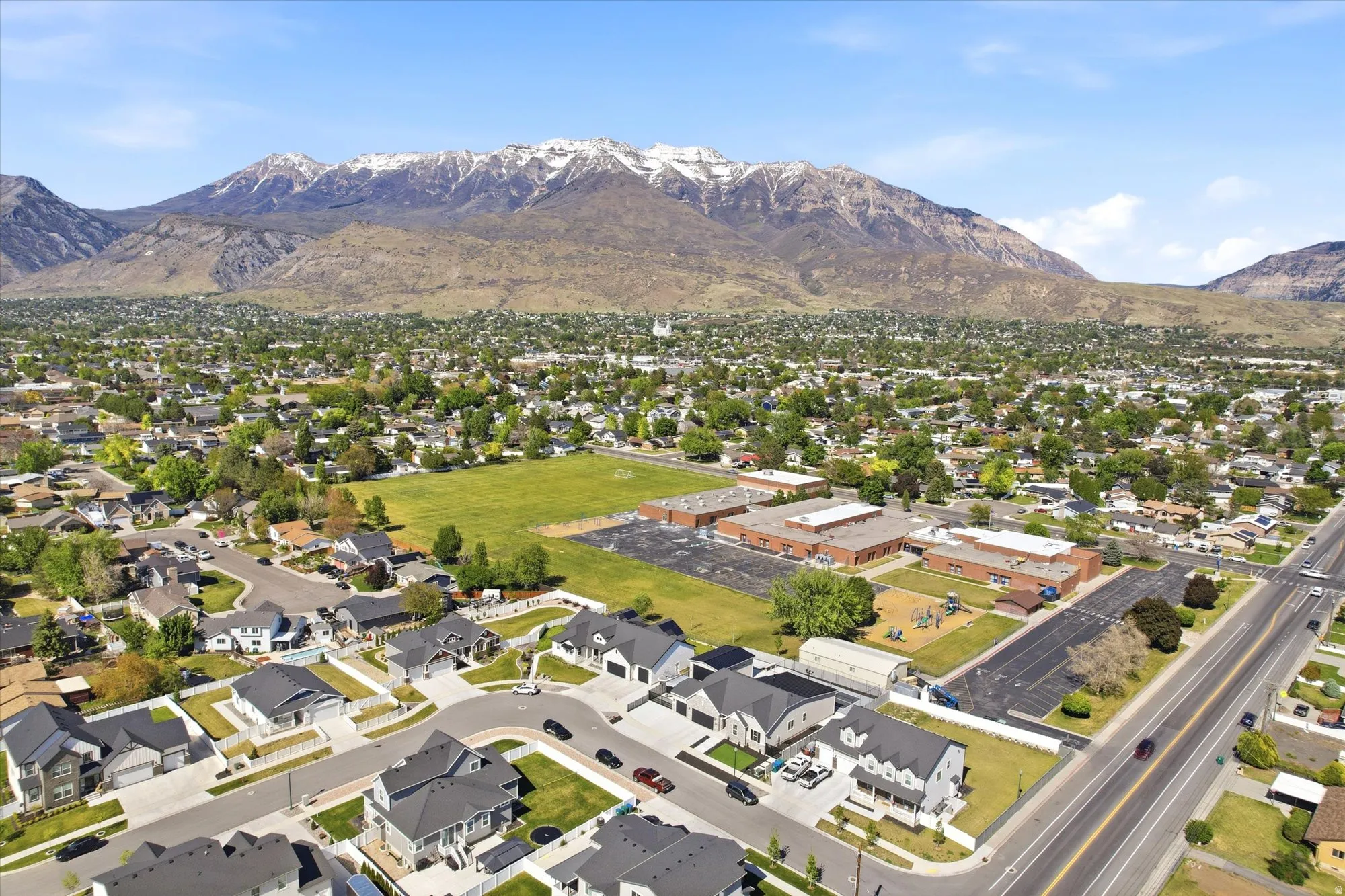 Expansive residential area featuring a distinct mountain backdrop with snow-capped peaks, a school building with a large adjacent green field, and a network of paved roads