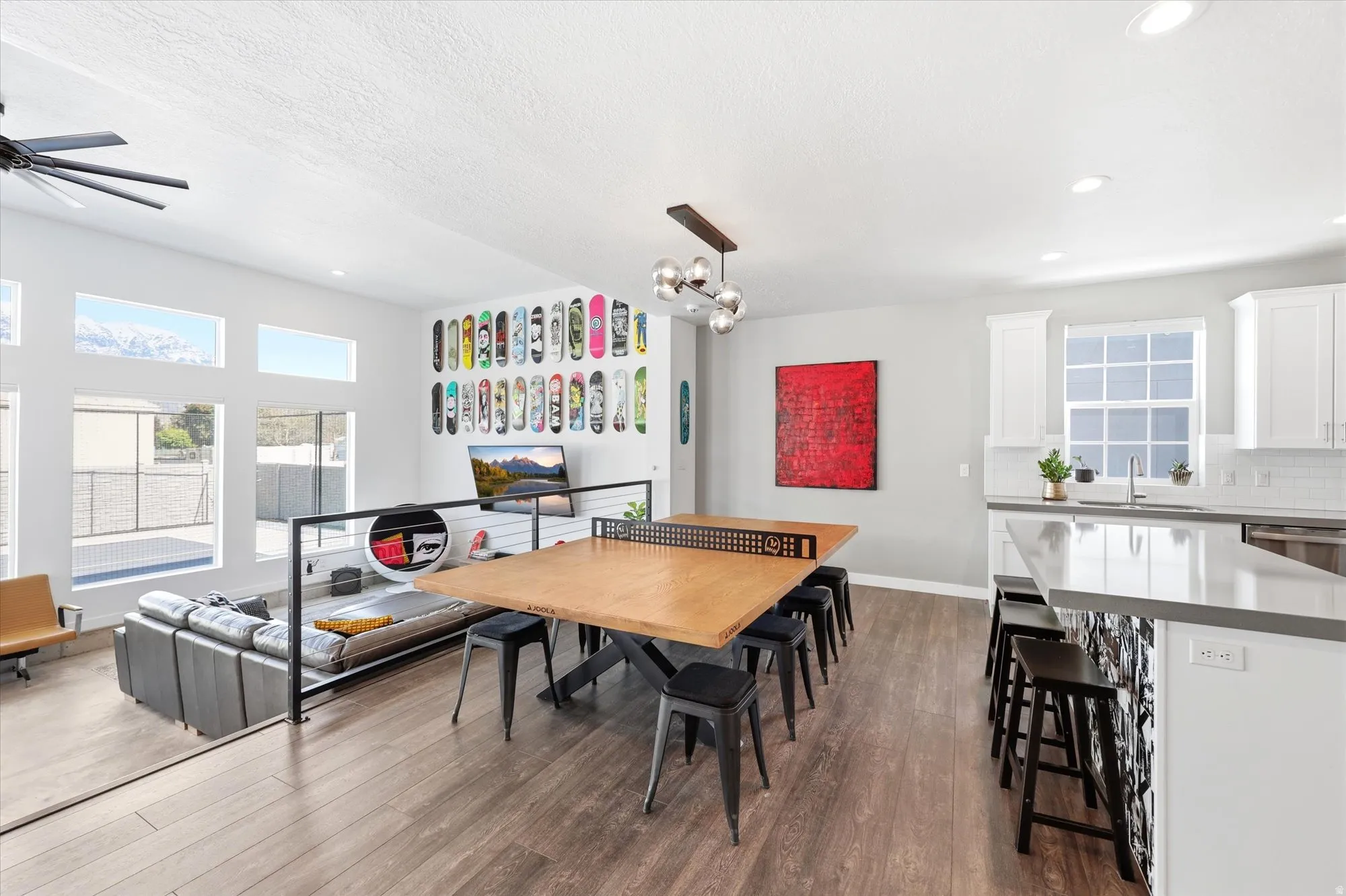 Open-concept living space featuring wood-finish flooring, a contemporary kitchen with white cabinetry and a light-toned countertop island, an integrated dining area with modern pendant lighting, and multiple large windows providing natural light