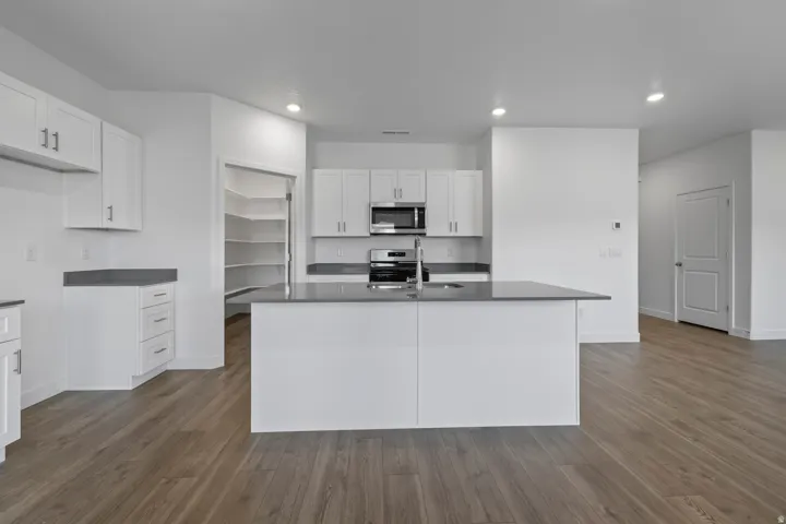 Modern kitchen featuring an island with an undermount sink, stainless steel appliances, white cabinetry, dark countertops, and wood-finish flooring