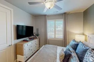 Bedroom featuring a ceiling fan with integrated lighting, window with plantation shutters and drapes, and a wall-mounted television