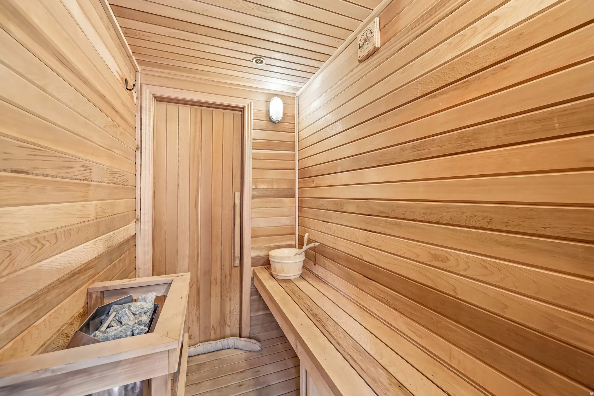 Wood-paneled sauna featuring a built-in bench, wood door, wall-mounted light fixture, and floor heater with rocks