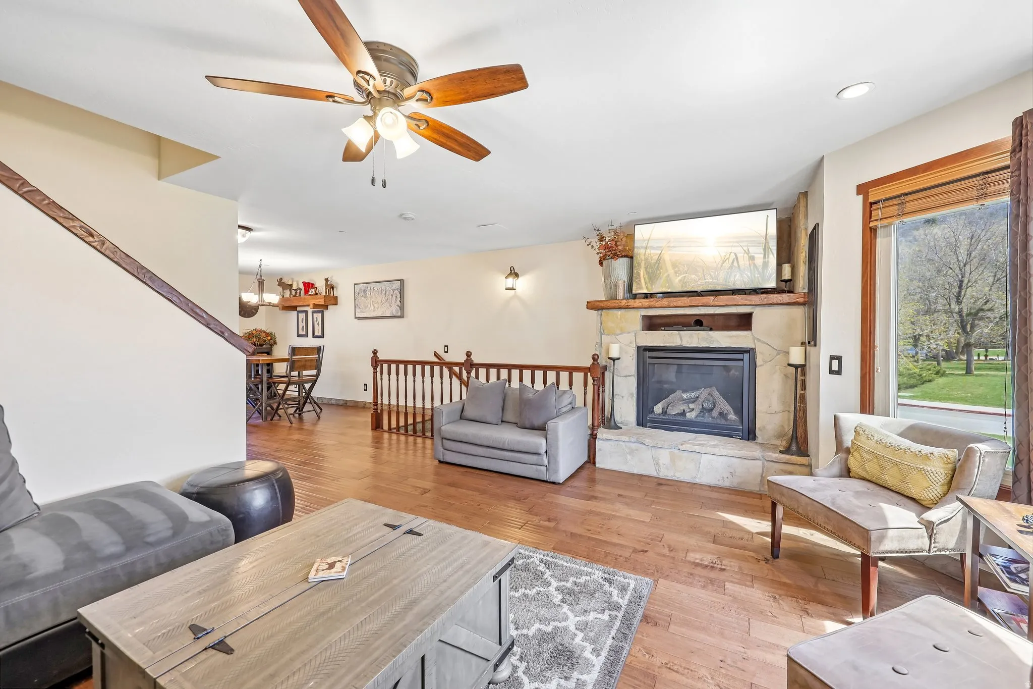 Living area featuring wood-finish flooring, a stone-surround fireplace with a wood mantle, and a ceiling fan with integrated lighting