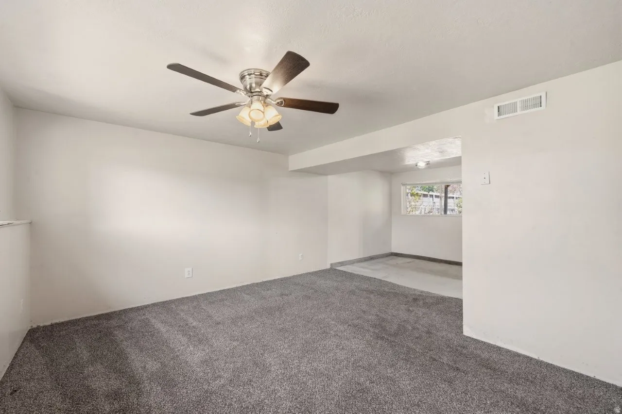 Spacious lower-level living room with carpet, ceiling fan, and an open sight line into the laundry and utility area. Great flex space with room for a sectional, gaming setup, or whatever your lifestyle needs.