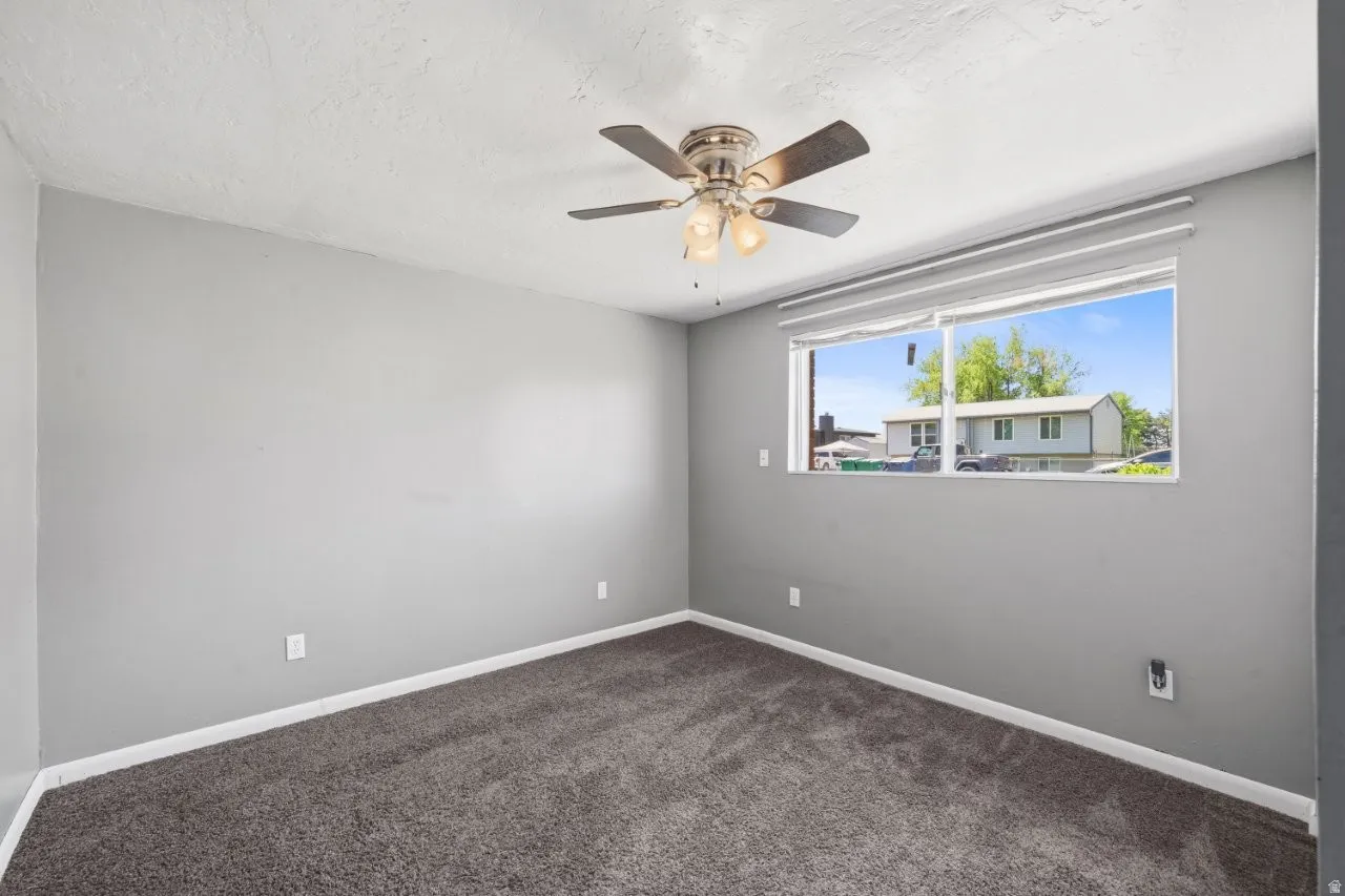 Updated bedroom with fresh gray paint, carpet, ceiling fan, and a bright window with a view of mature trees.