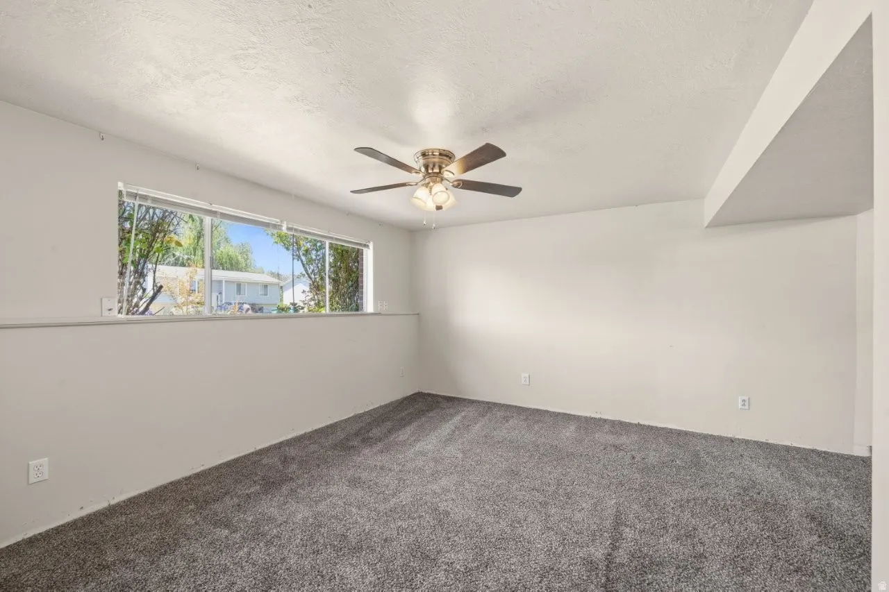 Generously sized lower-level room flooded with natural light from a wide picture window. Fresh white walls, carpet, and a ceiling fan make this a versatile space ready to become a family room, playroom, or additional living area.