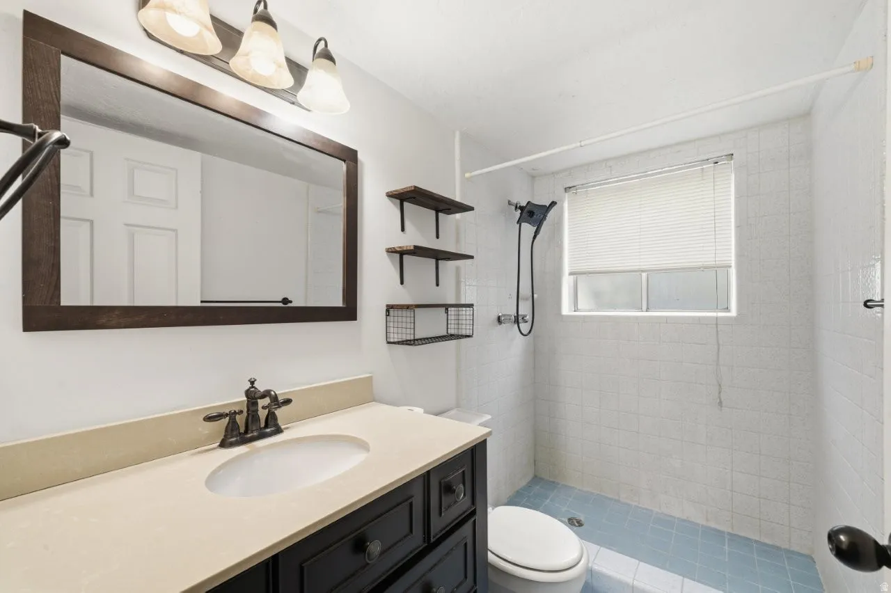 3/4 bathroom featuring a dark wood framed mirror, updated dark cabinetry vanity with stone-look countertop, oil-rubbed bronze fixtures, open tile shower with handheld showerhead, and rustic open shelving. Functional and full of character.