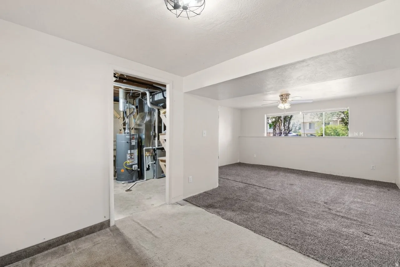 Lower-level utility room houses the water heater and furnace, keeping all the mechanicals accessible and organized. The adjacent carpeted living space with ceiling fan and window shows the flexibility this lower level offers.