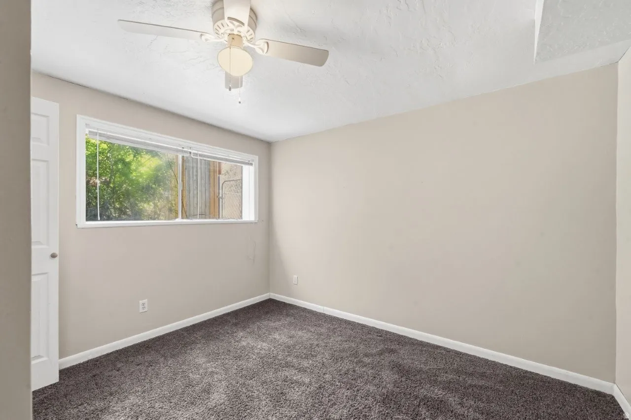 Cozy lower-level bedroom with carpet, ceiling fan, and a window overlooking the backyard and mature trees. Clean neutral walls and a private feel make this a great guest room, kids room, or home office.
