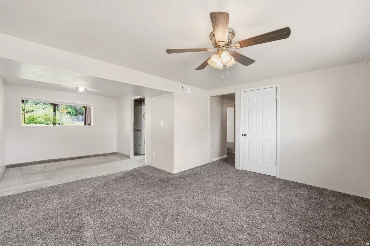 Another angle of the lower-level living space showing the open utility room with water heater and HVAC equipment, a window for natural light, and carpet throughout. Transparent and functional layout.