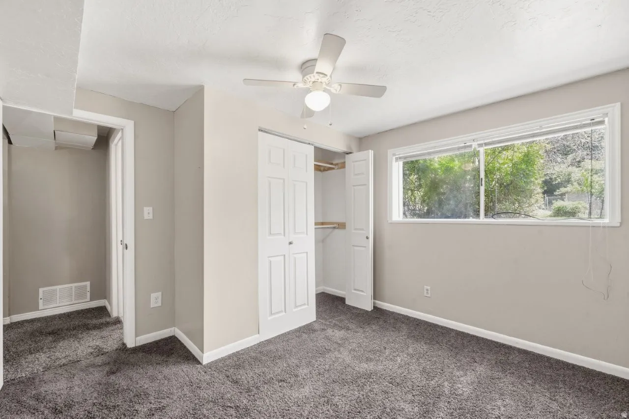 Good-sized lower-level bedroom with double door closet, ceiling fan, carpet, and a bright window with a green tree view.