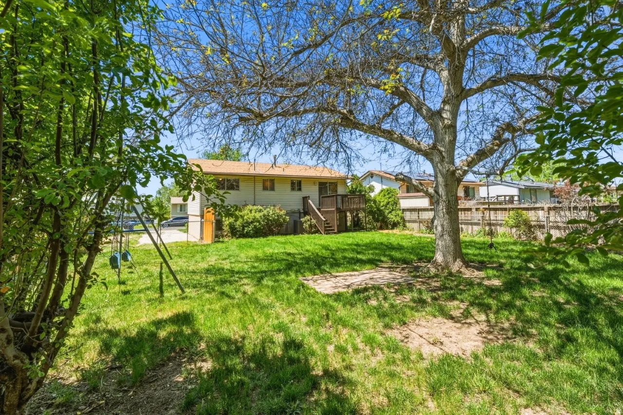 Charming rear view of the home framed by established trees and a swing set, with the back deck and staircase visible. This mature, private backyard is one of the best features of this property.
