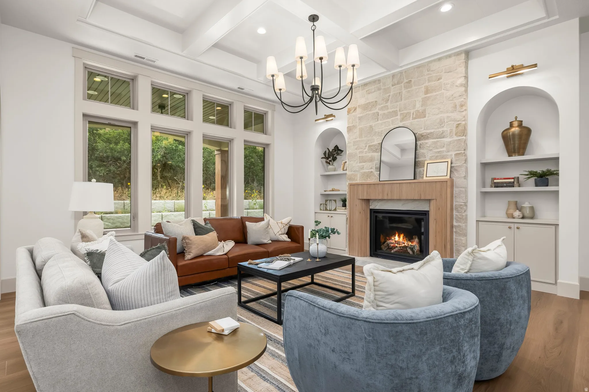 Coffered ceiling with a chandelier, stone-faced fireplace with wood surround, built-in arched shelving, large multi-pane windows, and wood-finish flooring