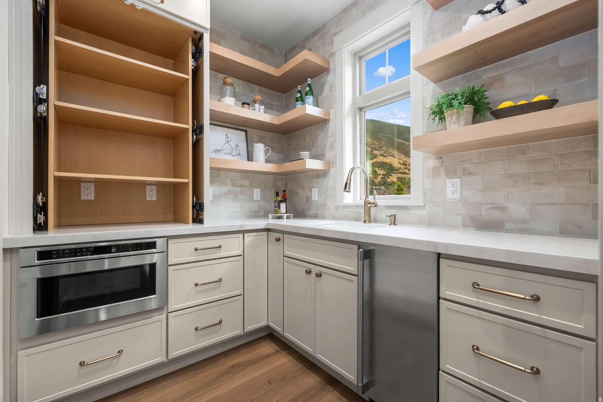Built-in microwave, under-counter sink, light wood-finish floating shelves, light-toned shaker-style cabinetry, and wood-finish flooring