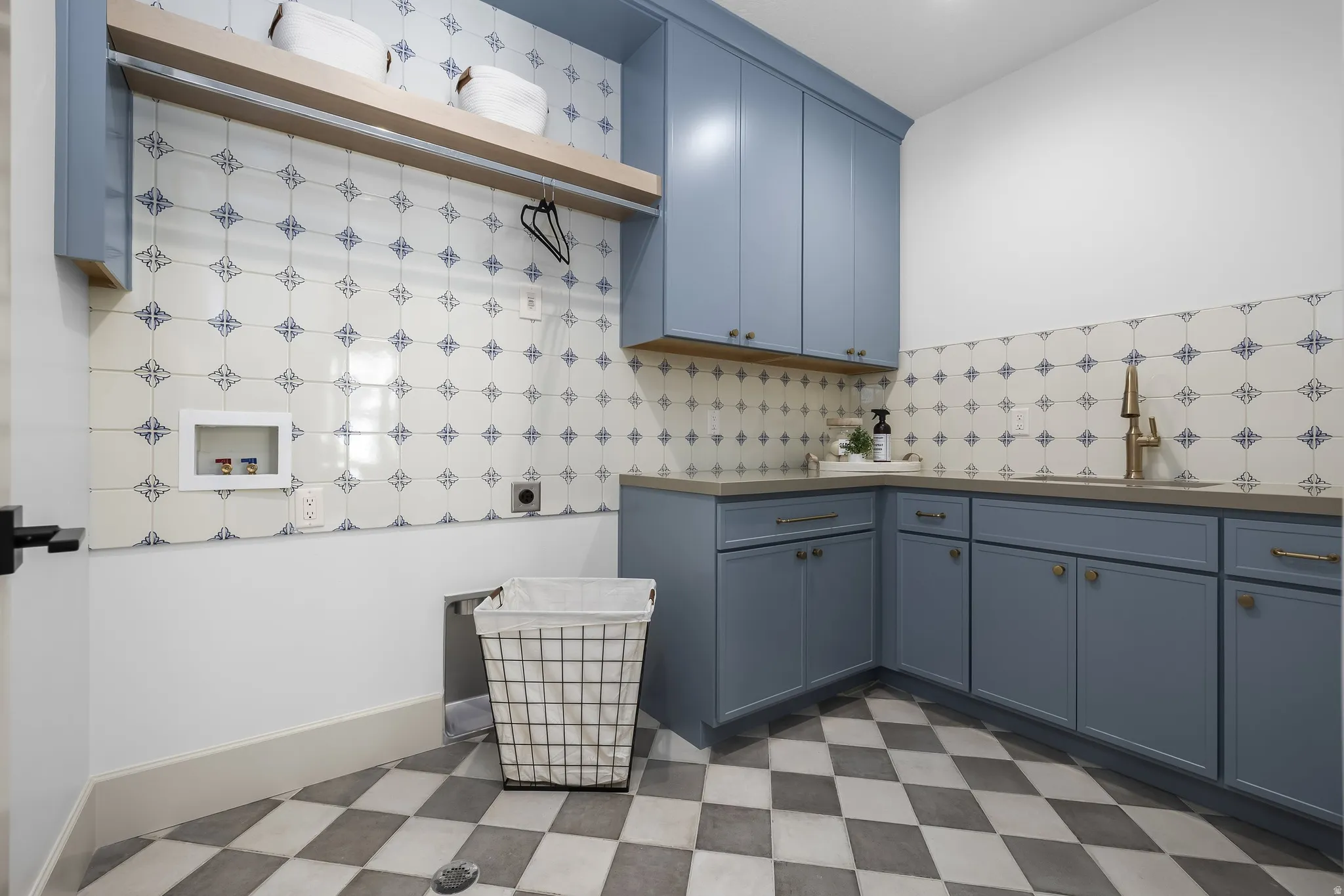 Laundry room featuring patterned tile flooring, blue cabinetry, a built-in sink with a gold-tone faucet, and a wall-mounted wood-finish shelf