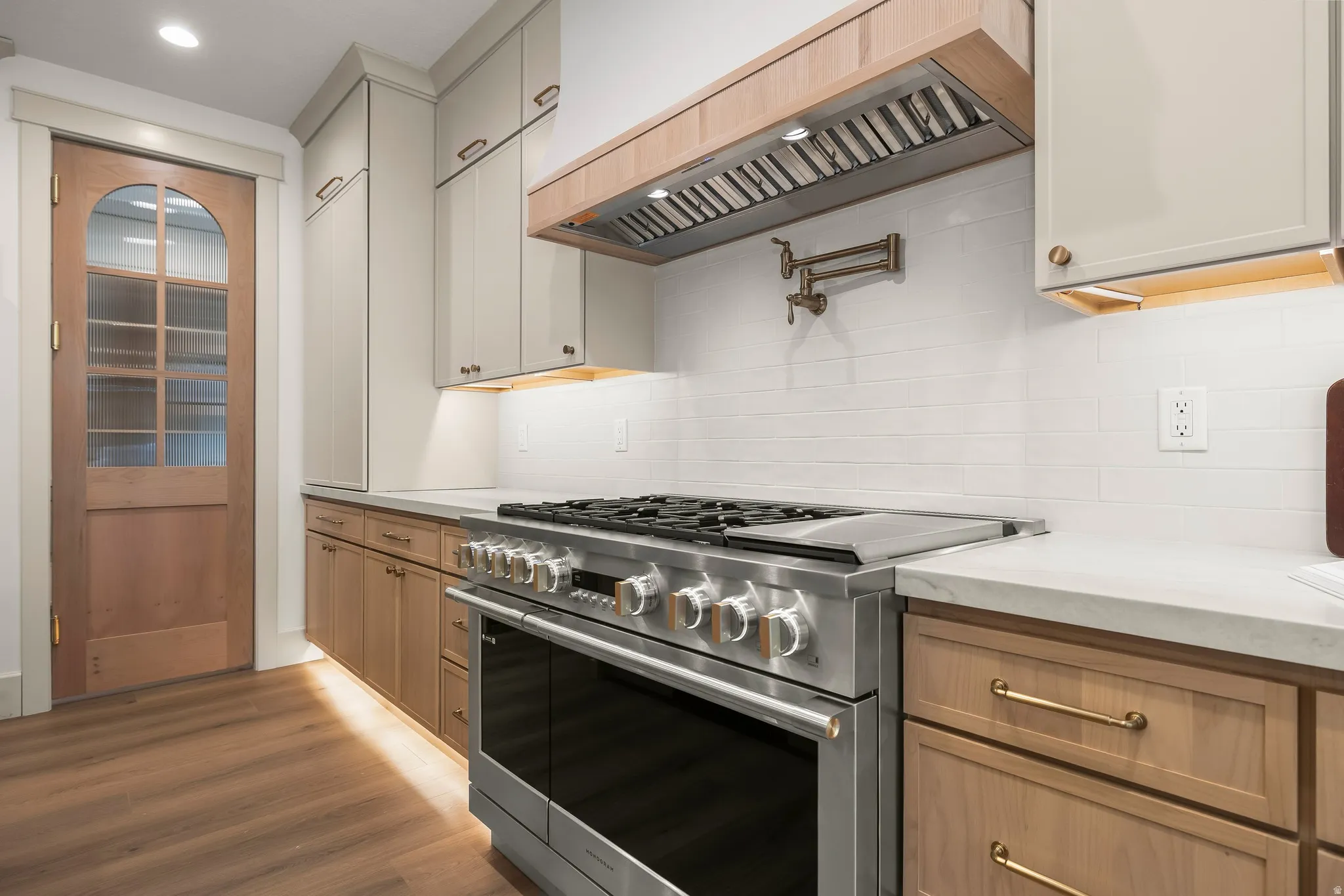 Stainless steel range with a pot filler, light wood-finish flooring, white subway tile backsplash, light-toned upper cabinetry, and natural wood lower cabinetry