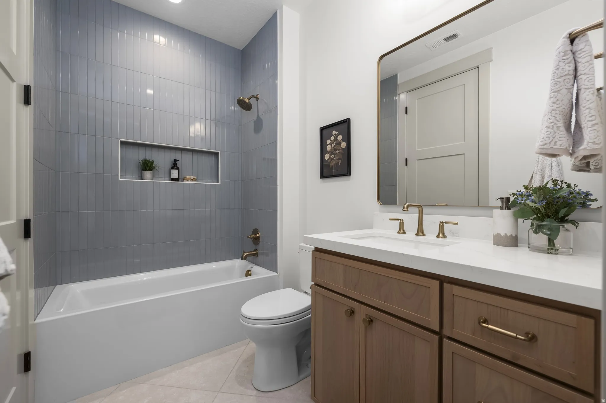 Bathroom featuring a tub/shower combination with blue vertical-stacked wall tile and an integrated niche, a single vanity with wood-finish cabinetry, a white countertop, and a framed mirror