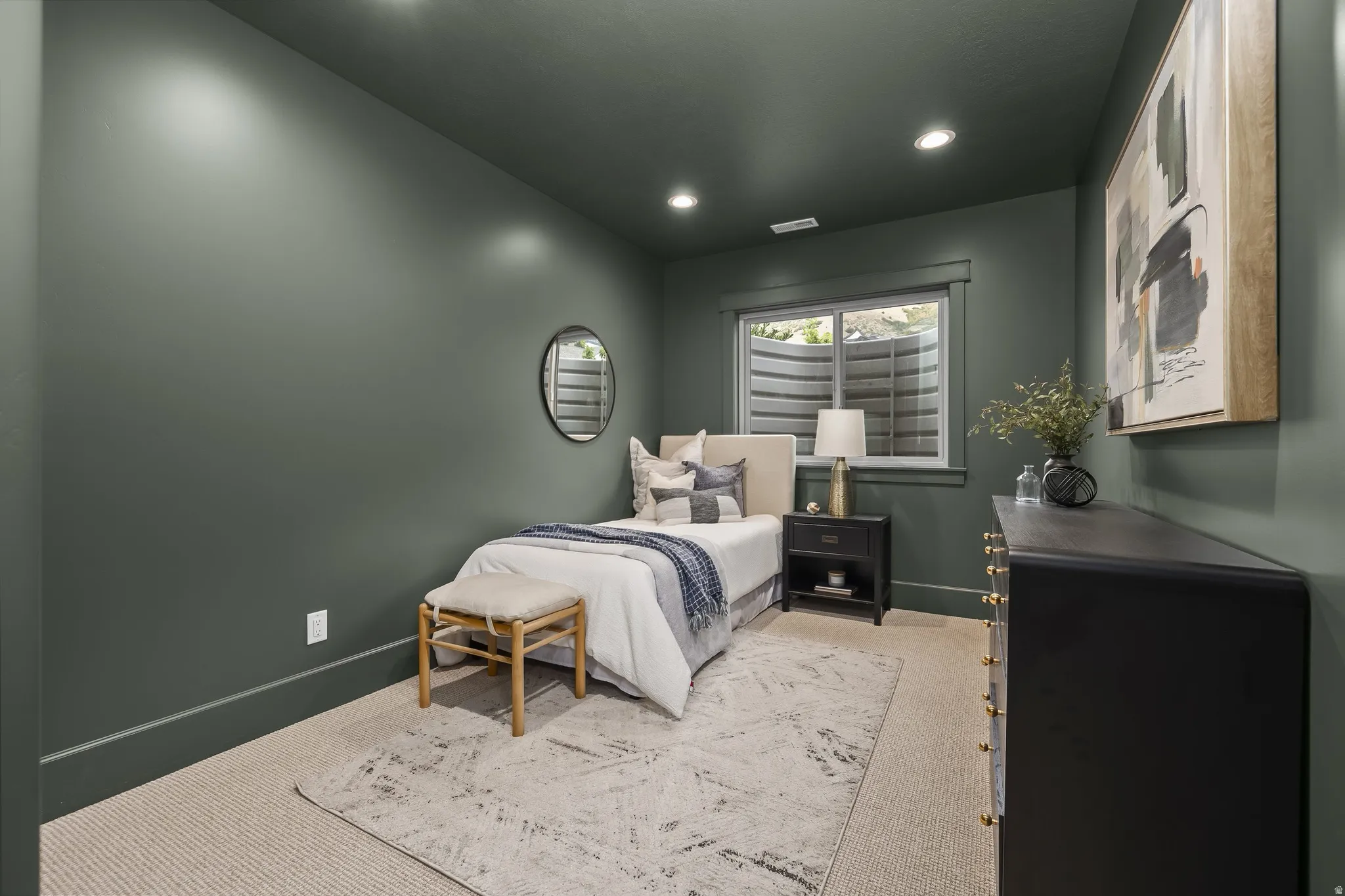 Recessed lighting, window with white trim, dark green wall color, light-colored carpeting, and a dark-toned dresser with gold-toned hardware