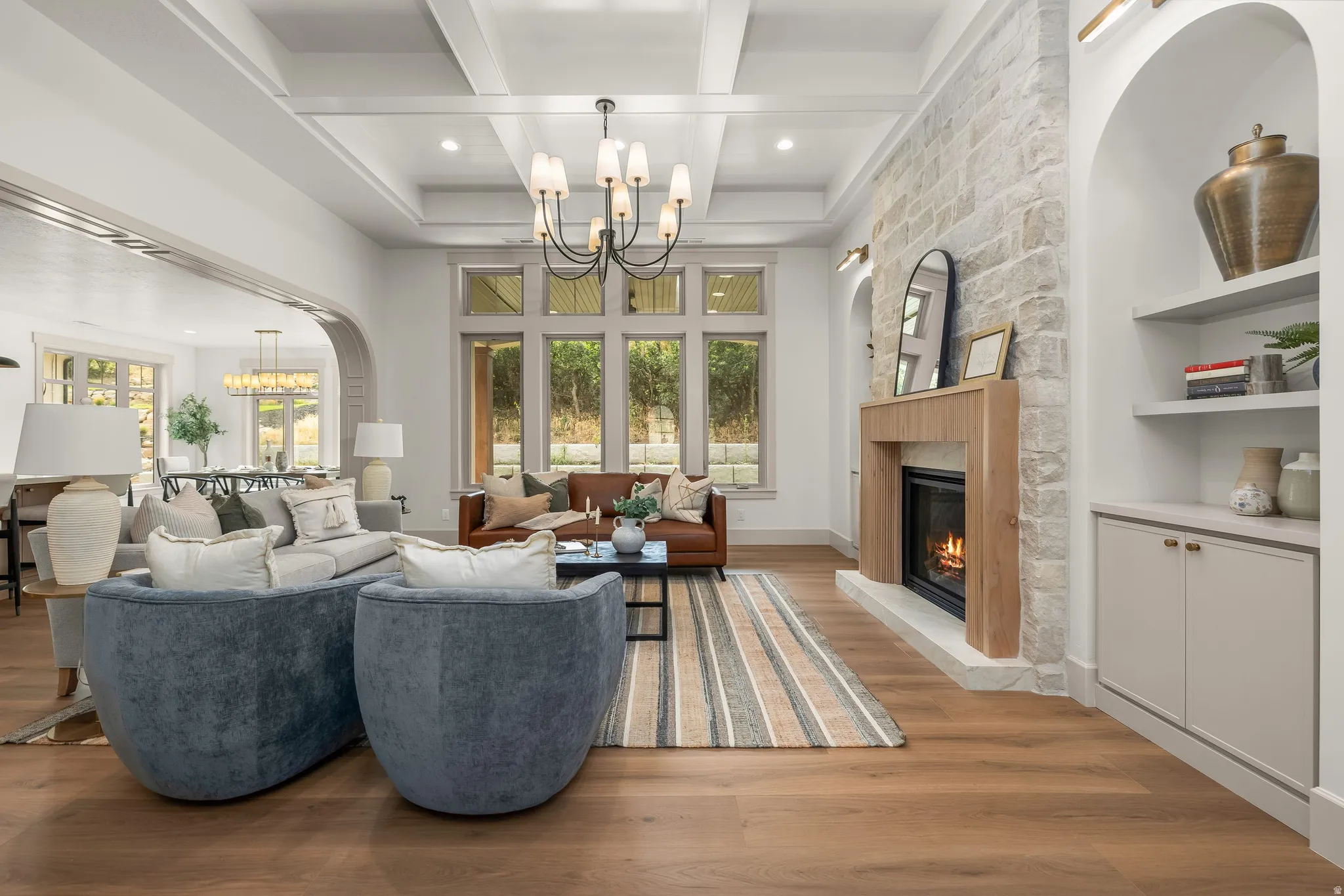 Wood-finish flooring throughout, coffered ceiling, stone-clad fireplace with wood mantel, large multi-pane windows, and built-in shelving with lower cabinetry