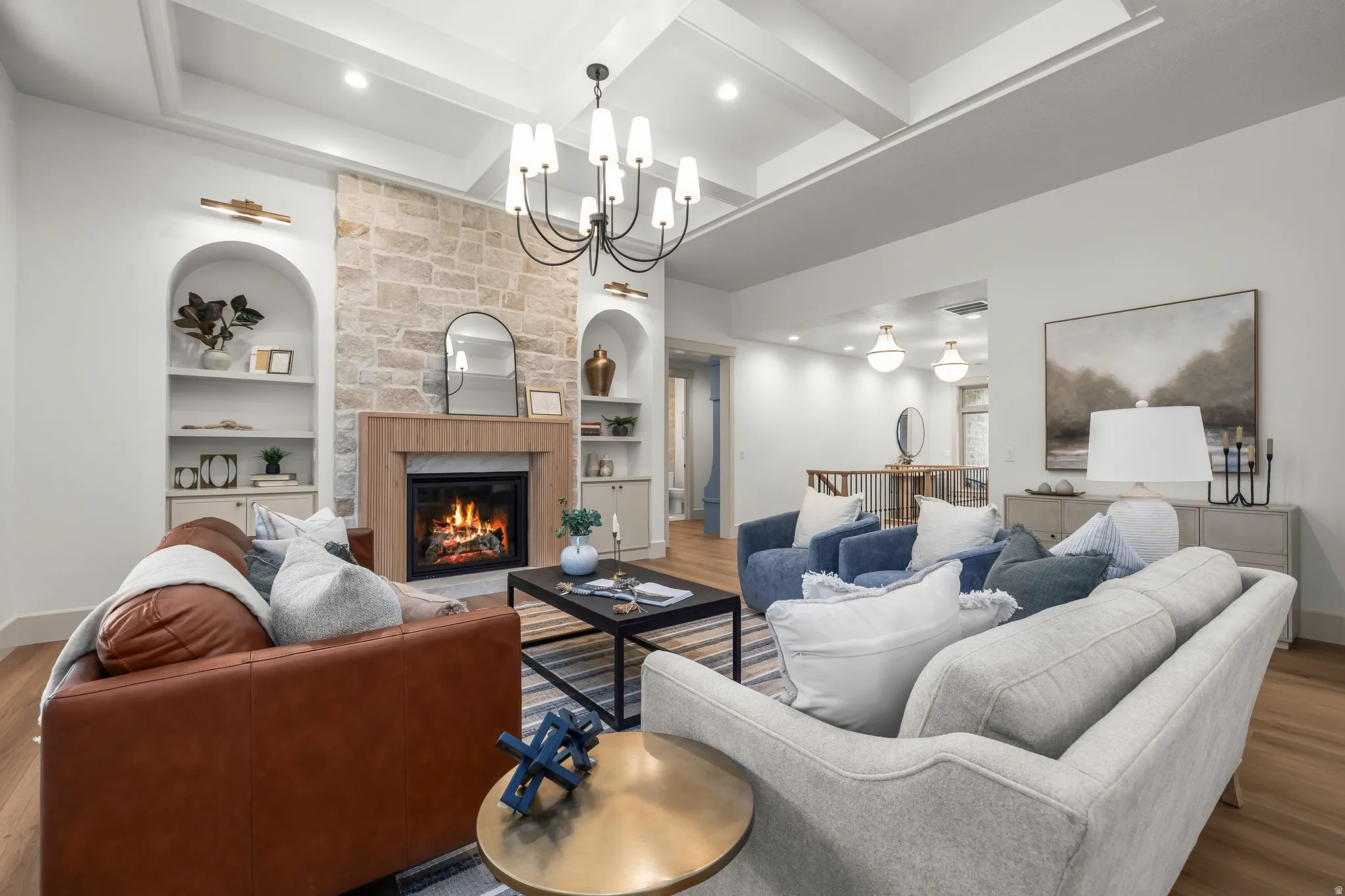 Vaulted ceiling with exposed beams, stone-clad fireplace with wood mantel, built-in arched shelving with cabinetry, wood-finish flooring, and a contemporary chandelier