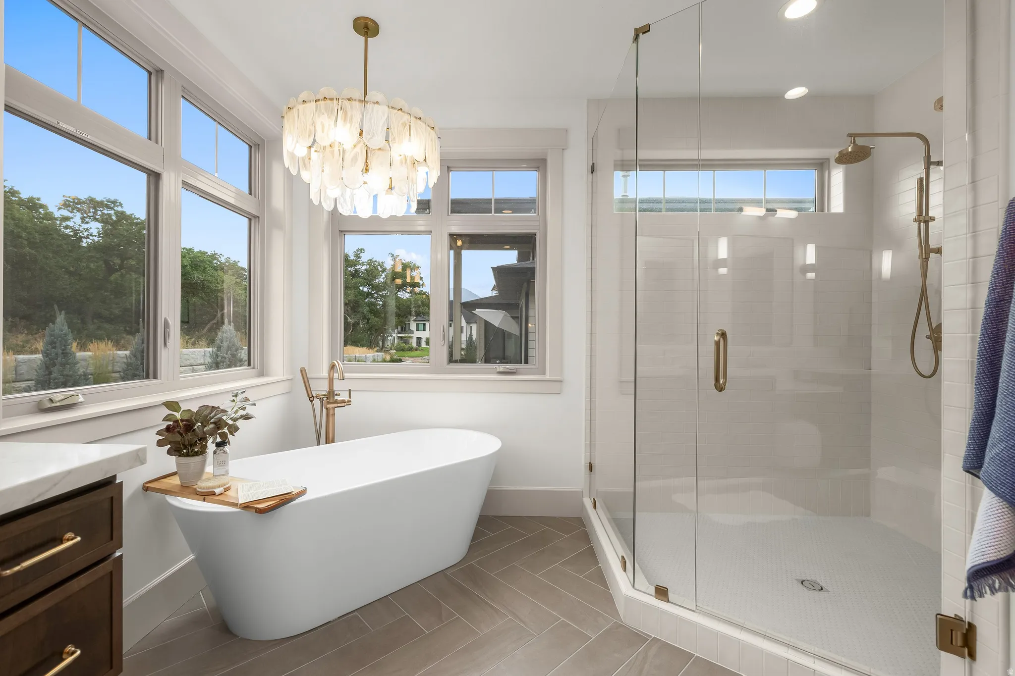 Freestanding soaking tub with a floor-mounted brass faucet, glass-enclosed shower with a rain shower head, dual pane windows, and a decorative chandelier