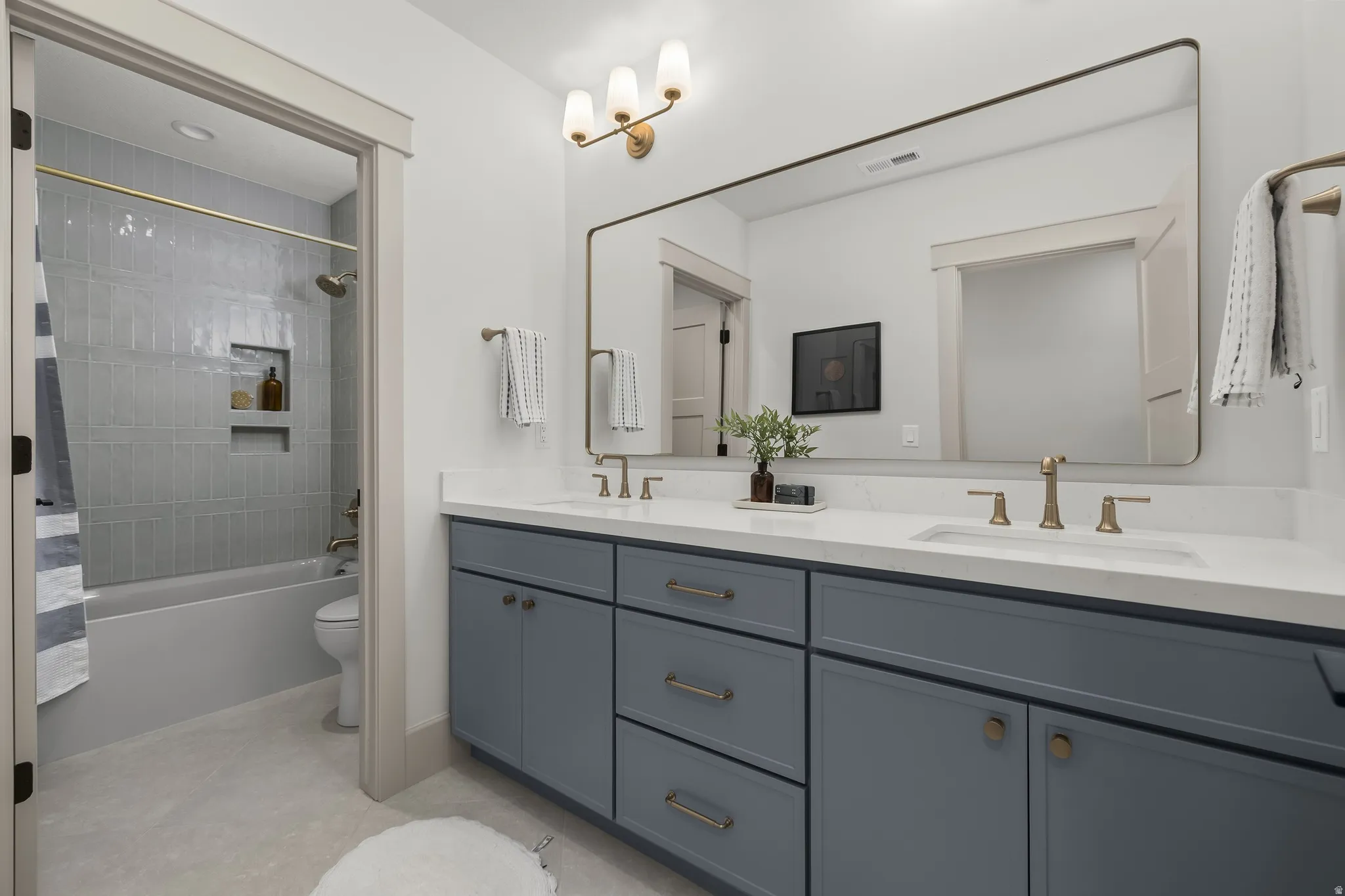 Double vanity with white countertop and integrated sinks, brushed gold fixtures, large frameless mirror, and a built-in shower niche