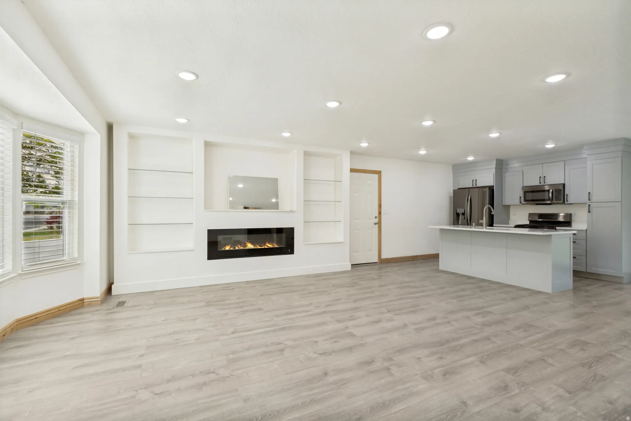 Open-concept living space with a contemporary electric fireplace featuring a built-in media center and shelving, wood-finish flooring, recessed lighting, a bay window with blinds, and a kitchen island with a sink and modern appliances